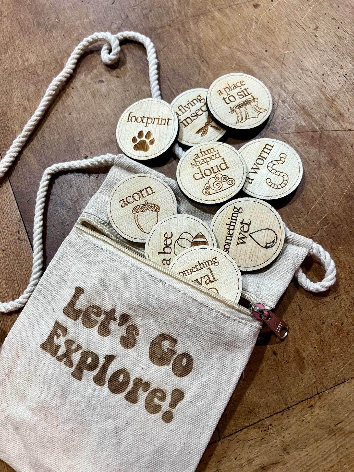 Wooden scavenger hunt coins spill from a drawstring bag that says "Let's Go Explore!".
