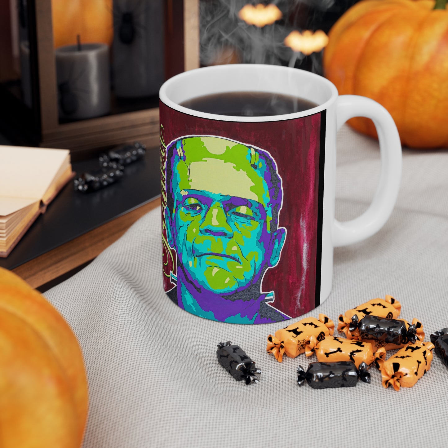 A white ceramic mug with a colorful Frankenstein monster design sits on a table with Halloween candy.