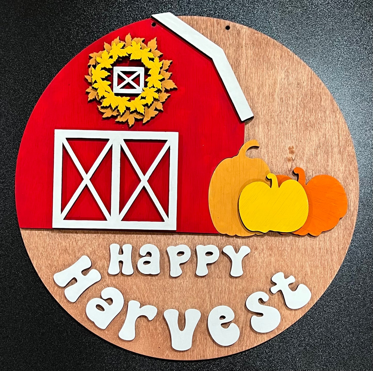 A round wooden sign with a red barn, fall leaves, pumpkins, and "Happy Harvest" text.