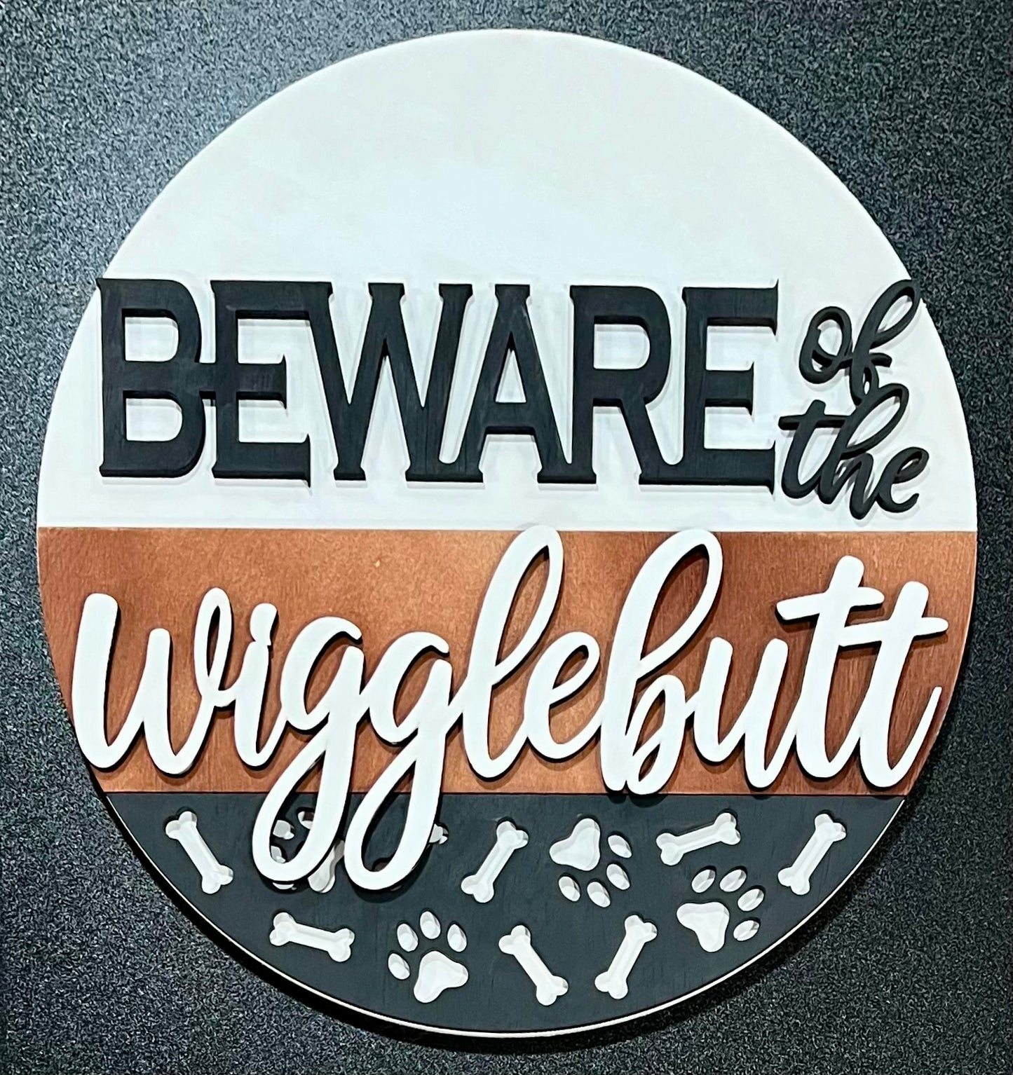 A round, unpainted wood sign reads "BEWARE of the wigglebutt" with paw prints and bones.