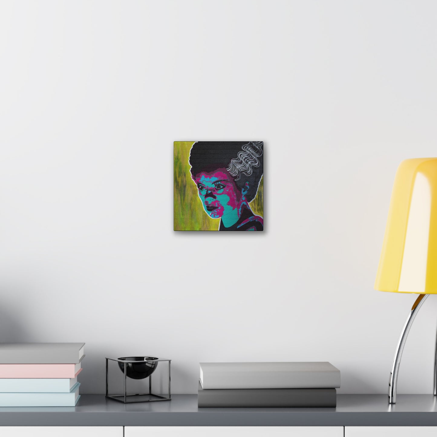 A canvas print of a woman with dark hair and bright pink and blue facial features hangs on a white wall.