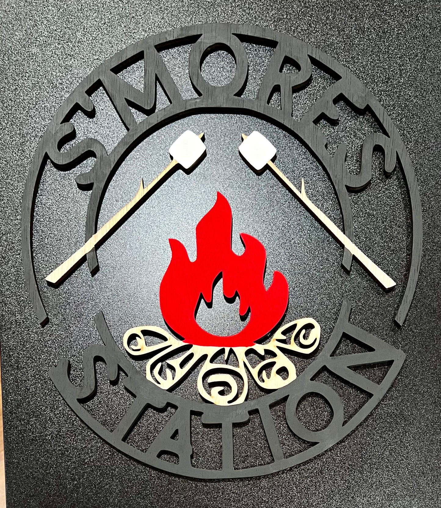 A black and red "S'mores Station" sign with two marshmallows on sticks over a campfire.