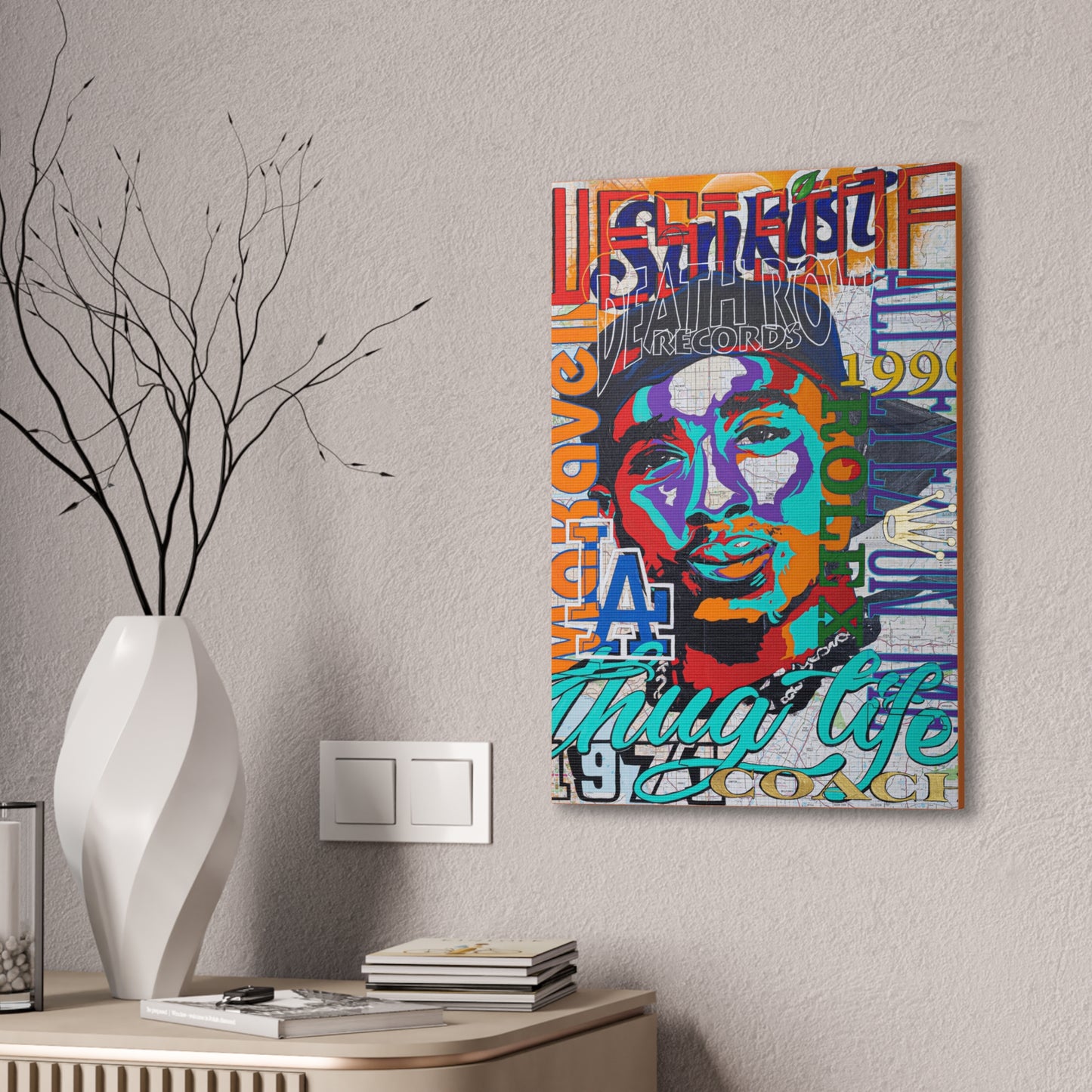 A colorful Tupac Shakur canvas print hangs on a textured wall above a white vase and books.