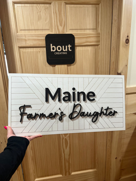 A white shiplap sign with "Maine Farmer's Daughter" in black script is held in front of a wooden door.