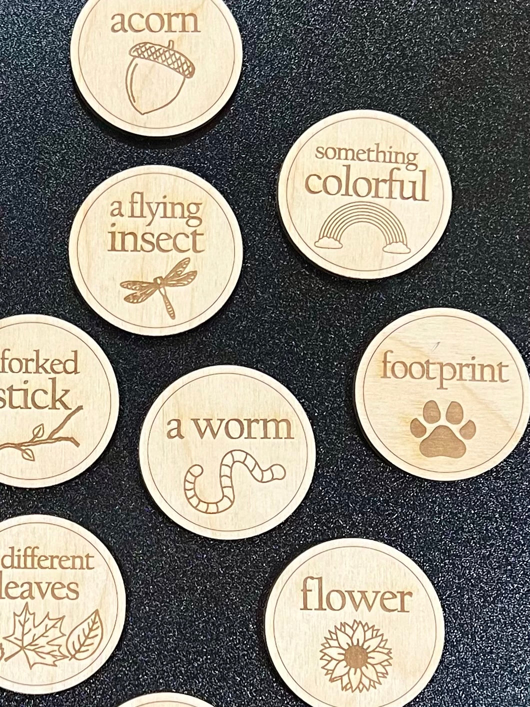 Nine round wooden tokens with engraved nature-themed words and illustrations are displayed.