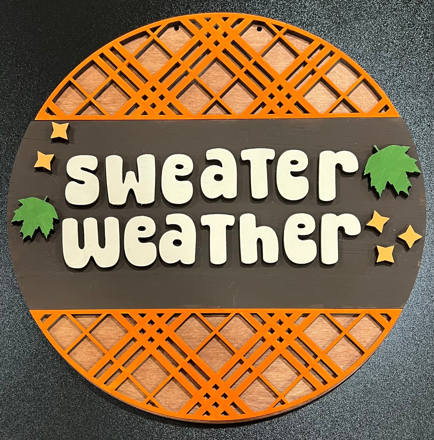 A round wooden sign with "Sweater Weather" in white letters, green leaves, and yellow stars.