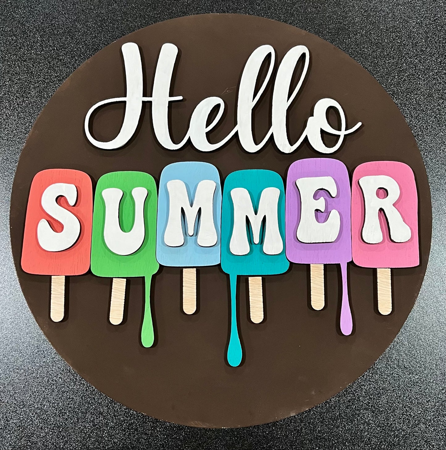 A round brown sign with "Hello Summer" in white letters, featuring colorful popsicle shapes.