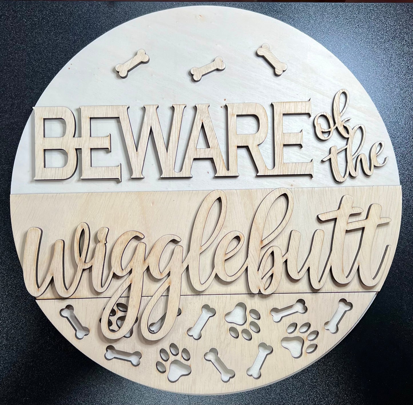 An unpainted round wooden sign reads "BEWARE of the wigglebutt" with bone and paw print cutouts.