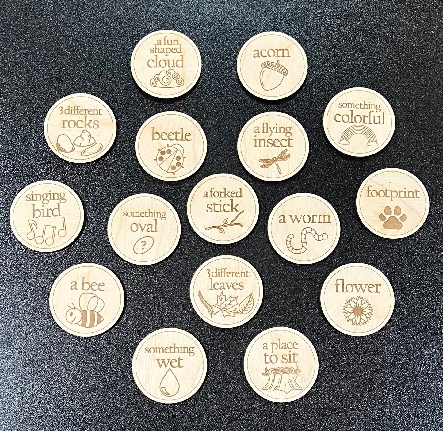 Seventeen wooden scavenger hunt coins with engraved nature-themed items are displayed on a dark surface.