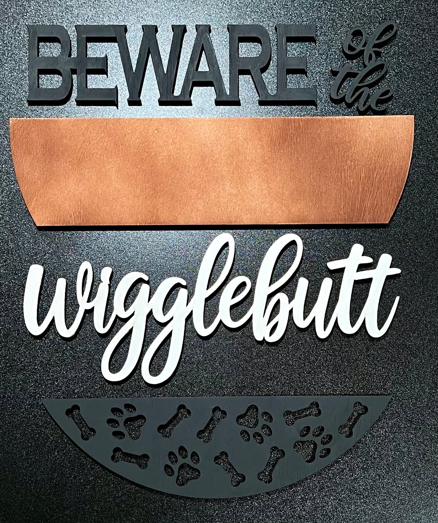 Unpainted wood sign reading "BEWARE of the wigglebutt" with paw prints and bones.