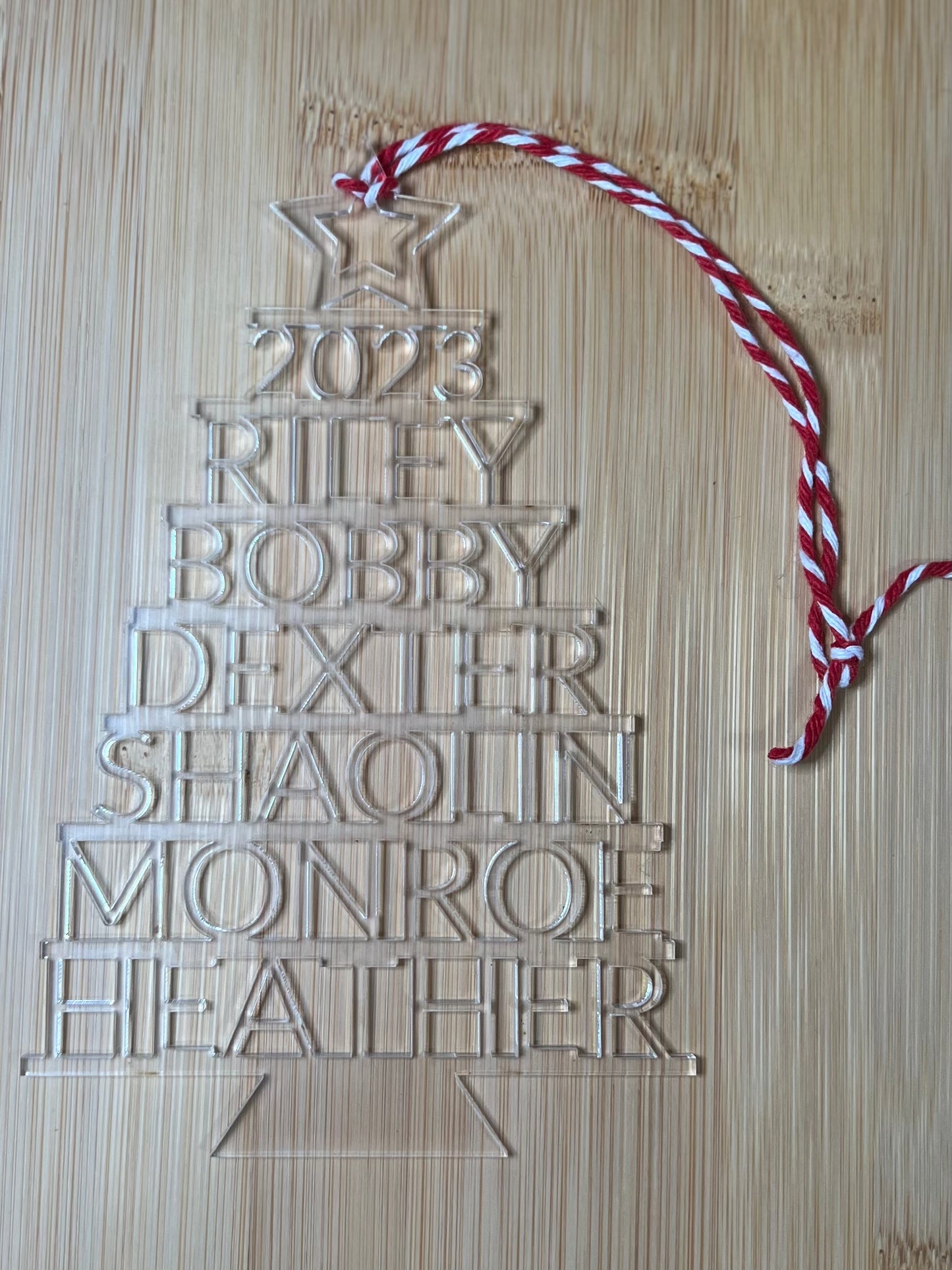 A clear acrylic Christmas tree ornament with "2023" and seven names stacked vertically.