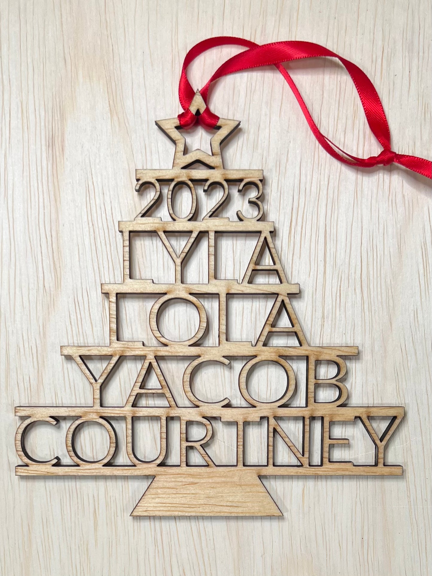 A wooden Christmas tree ornament with "2023," "LYLA," "LOLA," "JACOB," and "COURTNEY" stacked, tied with a red ribbon.