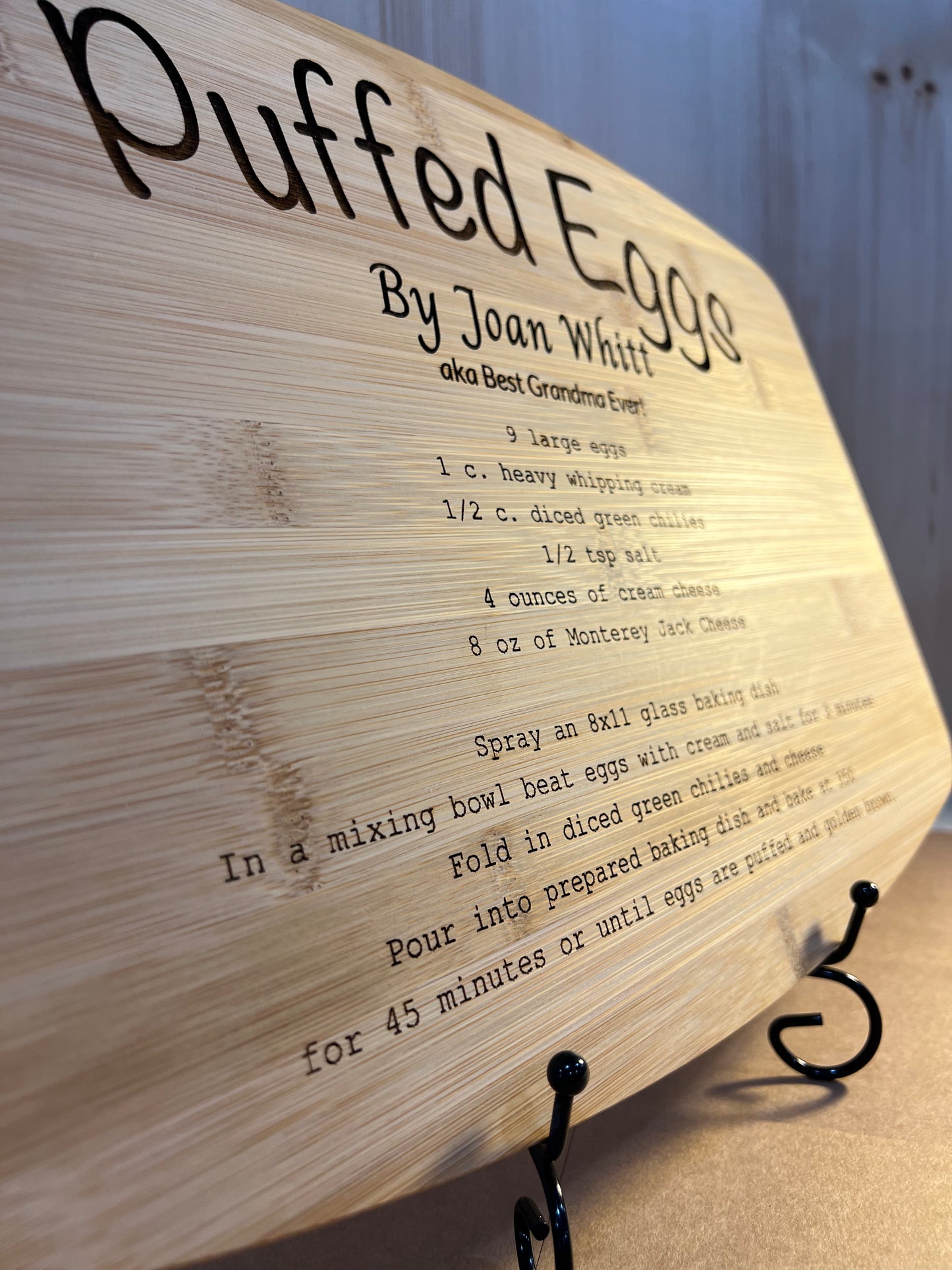 A bamboo cutting board with a "Puffed Eggs" recipe engraved on it, displayed on a stand.