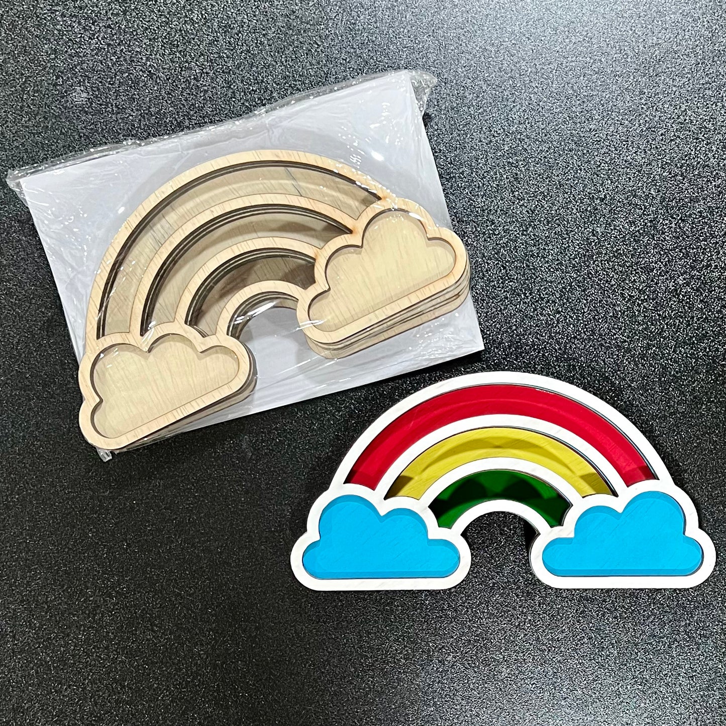 Two wooden rainbow-shaped craft kits, one unpainted and one painted, are on a dark surface.