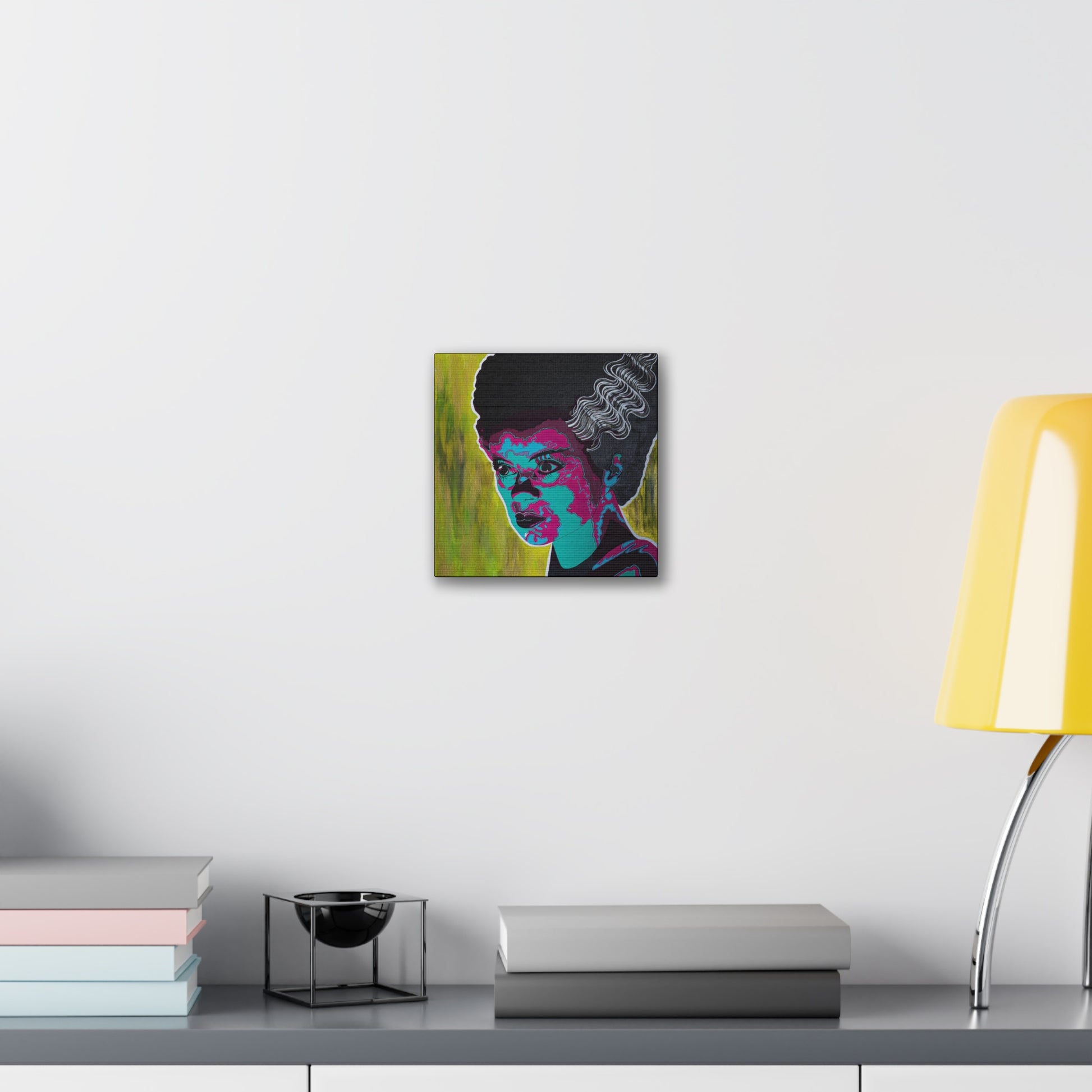 A canvas print of a woman with dark hair and bright pink and blue facial features hangs on a white wall.